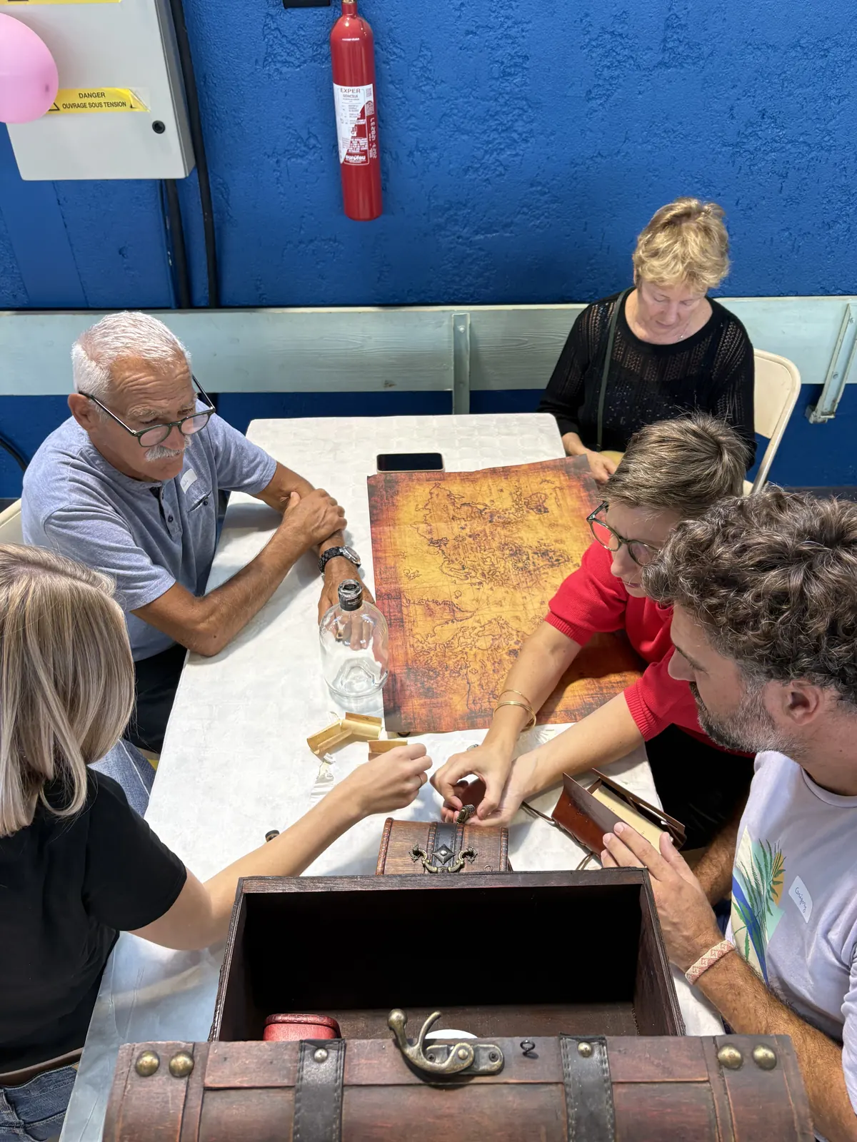 Cinq joueurs autour d'une table couverte d'une carte au trésor, penchés sur les objets sortis du coffre Pirate ouvert